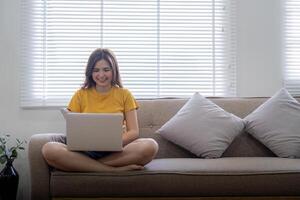 Jeune femme séance sur le canapé et travail sur projet, en train de regarder film sur portable du repos et content bavardage avec ami dans social réseau à Accueil photo