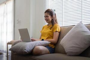 Jeune femme séance sur le canapé et travail sur projet, en train de regarder film sur portable du repos et content bavardage avec ami dans social réseau à Accueil photo