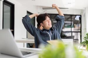 Jeune asiatique employé séance sur le chaise élongation lui-même et exercice pour relaxation tandis que travail difficile à Bureau photo