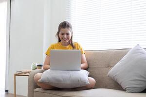 Jeune femme séance sur le canapé et travail sur projet, en train de regarder film sur portable du repos et content bavardage avec ami dans social réseau à Accueil photo