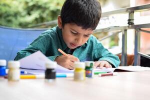 intelligent Indien peu garçon effectuer pouce La peinture avec différent coloré l'eau Couleur trousse pendant le été les vacances, mignonne Indien enfant Faire coloré pouce La peinture dessin sur en bois table photo