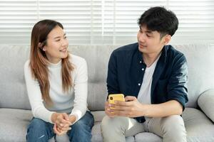 asiatique couple souriant et Heureusement utilisation téléphone intelligent à en ligne achats. mari prêt à Payer anniversaire présent pour magnifique épouse. trouver hébergement en ligne pour voyage de noces, app en ligne, livre vol billet photo