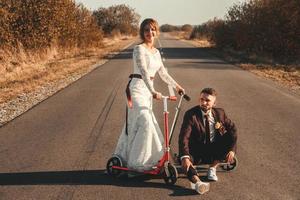 couple de mariage souriant chevauchant un scooter le long de la route à l'extérieur de la ville au coucher du soleil. photo