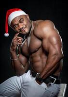 taille en haut portrait de le africain américain bodybuilder homme avec torse nu musclé torse. Masculin portant Noël chapeau isolé sur noir Contexte photo