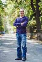 homme avec gris cheveux Extérieur dans parc. non veste. bleu chandail. lunettes. barbu sur de soi homme d'affaire. flou Contexte. photo