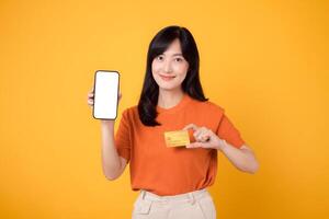 joyeux asiatique femme dans sa 30s, révélateur Vide téléphone intelligent écran et crédit carte sur vibrant Jaune Contexte. rapide en ligne Paiement achats. photo