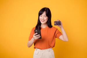 enthousiaste asiatique femme dans sa 30s, en utilisant téléphone intelligent et montrant crédit carte sur Jaune Contexte. rapide en ligne achats. photo