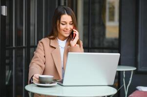 Jeune content affaires Dame regards à téléphone intelligent écran avec à pleines dents sourire tandis que séance à l'extérieur sur terrasse avec portable et café, travail à distance en ligne vérification rapports et avoir prêt pour réunion photo