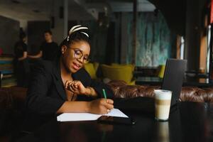 magnifique afro américain fille dans décontractée vêtements est en utilisant une portable et souriant tandis que séance dans café photo