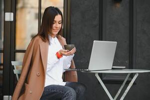 Jeune pigiste femme profiter café tandis que travail en plein air sur une portable ordinateur connecté à Publique Wifi. élégant étudiant fille en train d'étudier en ligne dans ville. photo