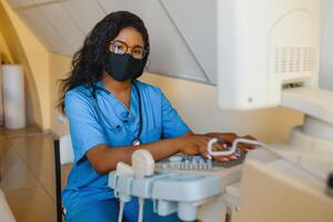 Jeune femelle Afro-américain médecin travail sur moderne ultrason équipement. opérateur de ultrason balayage machine séance et à la recherche à le moniteur, attendre pour patient. photo