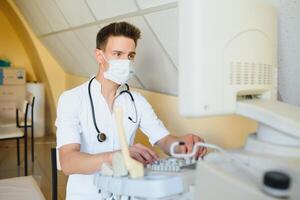 Masculin médecin dans masque avec ultrason équipement regards à le moniteur dans le clinique Bureau photo