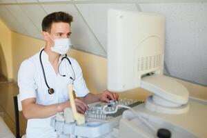 Masculin médecin dans masque avec ultrason équipement regards à le moniteur dans le clinique Bureau photo