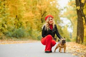 une élégant Jeune fille avec longue lumière cheveux dans ensoleillé des lunettes va pour une marcher avec une peu milieu chienchien une carlin par le français bouledogue dans une parc dans printemps dans l'automne photo