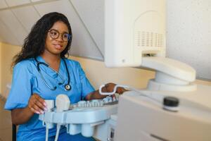 souriant africain femme médecin avec ultrason scanner dans main, travail sur moderne ultrason balayage machine dans lumière pièce dans clinique. portrait de 4d ultrason balayage machine opérateur photo