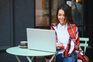 Jeune pigiste femme profiter café tandis que travail en plein air sur une portable ordinateur connecté à Publique Wifi. élégant étudiant fille en train d'étudier en ligne dans ville. photo