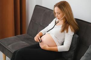 le mère est en utilisant stéthoscope le médecin écoute à le du son de le bébé dans le estomac. asiatique Enceinte femme en utilisant stéthoscope écoute sa bébé sur canapé. photo