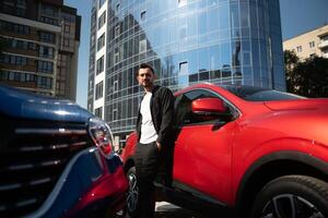 un attrayant homme avec une barbe des stands suivant à le sien voiture dans le parking parcelle. il est réfléchi, et regards loin, Quelqu'un est en attendant. été journée photo