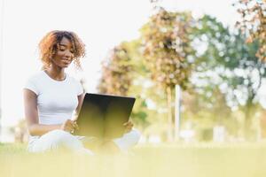 Humain et La technologie concept. charmant africain femme avec court lutin coiffure profiter ensoleillé temps, séance sur le pelouse dans de face de portable ordinateur dans le Publique jardin, attendre pour sa ami photo