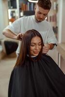 une content Jeune femme est assis dans une chaise couvert avec une cap tandis que une cheveux Maître coupes sa cheveux photo