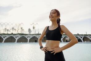 souriant femme dans tenue de sport avoir une du repos après faire des exercices à l'extérieur permanent sur bâtiment Contexte photo