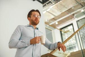 homme d'affaire permanent avec tasse de café dans Bureau escaliers pendant Pause temps et à la recherche à côté photo