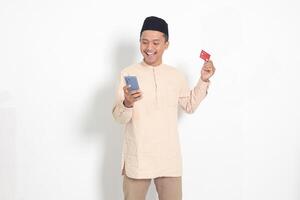 portrait de attrayant asiatique musulman homme dans koko chemise avec calotte en portant une mobile téléphone et en présentant crédit carte. isolé image sur blanc Contexte photo