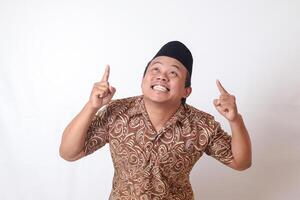 portrait de excité asiatique homme portant batik chemise et Songkok souriant et à la recherche à le caméra montrer du doigt avec deux mains et les doigts à en haut. isolé image sur gris Contexte photo