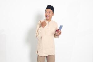 portrait de attrayant asiatique musulman homme dans koko chemise avec calotte en portant une mobile téléphone et crédit carte tandis que montrer du doigt avec doigt à le côté. isolé image sur blanc Contexte photo