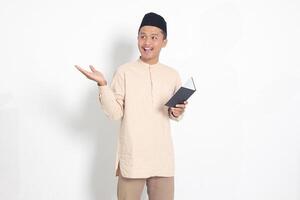 portrait de excité musulman homme dans koko chemise avec peci en train de lire une carnet. content asiatique gars montrer du doigt à le côté avec doigt. isolé image sur blanc Contexte photo