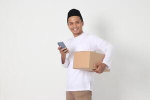 portrait de excité asiatique musulman homme dans koko chemise avec peci porter papier carton boîte tandis que en portant mobile téléphone. Aller Accueil pour eid moubarak. isolé image sur blanc Contexte photo
