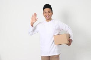 portrait de excité asiatique musulman homme dans koko chemise avec peci en disant Salut, salutation à Quelqu'un tandis que porter papier carton boîte. Aller Accueil pour eid moubarak. isolé image sur blanc Contexte photo