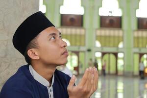 portrait de religieux asiatique homme dans musulman chemise et noir casquette prier avec main élevé à l'intérieur le Publique mosquée photo