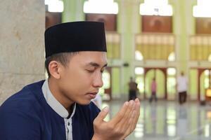 portrait de religieux asiatique homme dans musulman chemise et noir casquette prier avec main élevé à l'intérieur le Publique mosquée photo