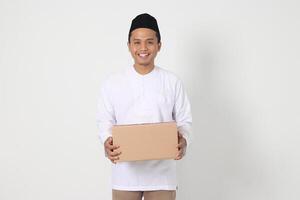 portrait de excité asiatique musulman homme dans koko chemise avec peci porter papier carton boîte. Aller Accueil pour eid moubarak. isolé image sur blanc Contexte photo