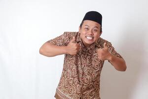 portrait de excité asiatique homme portant batik chemise et Songkok souriant et à la recherche à caméra, fabrication les pouces en haut main geste. isolé image sur gris Contexte photo