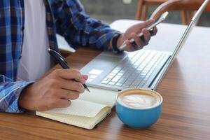 proche en haut de Masculin main l'écriture sur Remarque livre avec stylo, montrant Vide écran de portable et téléphone intelligent. travail dans café concept avec une tasse de café photo