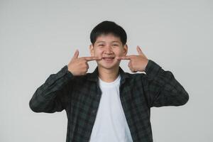 content asiatique adolescent garçon montrer du doigt à croisillons. souriant asiatique adolescent homme montrer du doigt à le sien dentaire un appareil dentaire sur une lumière gris Contexte. portrait Beau homme dans studio concept. photo