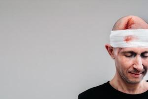 contre une gris Contexte des stands une battu et battu homme dans une noir T-shirt avec le sien tête enveloppé dans une bandage photo