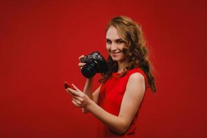 une souriant Jeune femme avec ondulé cheveux détient une fraise et photographies il, en portant une délicieux Frais fraise sur une brillant rouge Contexte photo