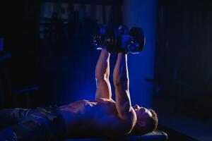 Beau Puissance athlétique homme bodybuilder Faire des exercices avec haltère. aptitude musclé corps sur noir et bleu Contexte. aptitude concept. photo