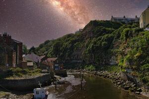 pittoresque côtier village à nuit avec illuminé laiteux façon galaxie au-dessus de, serein rivière, et traditionnel Maisons dans Staithes, Angleterre. photo