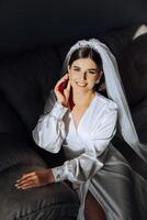 portrait de un incroyablement magnifique fille la mariée dans une blanc peignoir dans le chambre à coucher, le la mariée pose dans le Matin avant le mariage cérémonie. photo
