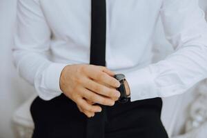 une fermer de une tondu Cadre de une homme met sur une regarder avec une cuir ceinture, est habillé dans une élégant costume, une blanc chemise, porte une or anneau. photo