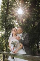 une Jeune la mariée et jeune marié tendrement embrasse dans le des rayons de le l'automne Soleil. soumissionner et magnifique Jeune fille la mariée. une homme baisers le sien bien-aimé. contre le Contexte de une magnifique jardin photo