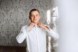 une homme dans une blanc chemise des stands par le fenêtre dans le pièce et attache le boutons sur le sien collier et manches. regarder sur main. élégant affaires portrait de une homme, fermer photo. le jeune marié est en train de préparer. photo