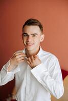 une homme dans une blanc chemise des stands par le fenêtre dans le pièce et attache le boutons sur le sien collier et manches. regarder sur main. élégant affaires portrait de une homme, fermer photo. le jeune marié est en train de préparer. photo