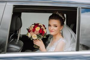 le la mariée est séance dans une noir voiture avec une bouquet de fleurs dans sa mains et regards en dehors de le auto. portrait de une plutôt timide la mariée dans une auto. mariée, sourire, émotions. photo