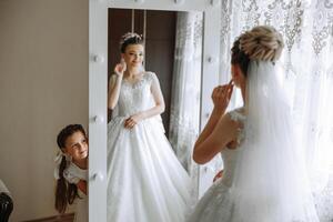 portrait de le la mariée avec sa peu sœur dans le chambre. le peu sœur de le la mariée regards à le la mariée de derrière le miroir. le la mariée est en train de préparer pour le mariage photo