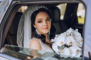 le la mariée est séance dans une noir voiture avec une bouquet de fleurs dans sa mains et regards en dehors de le auto. portrait de une plutôt timide la mariée dans une auto. mariée, sourire, émotions. photo